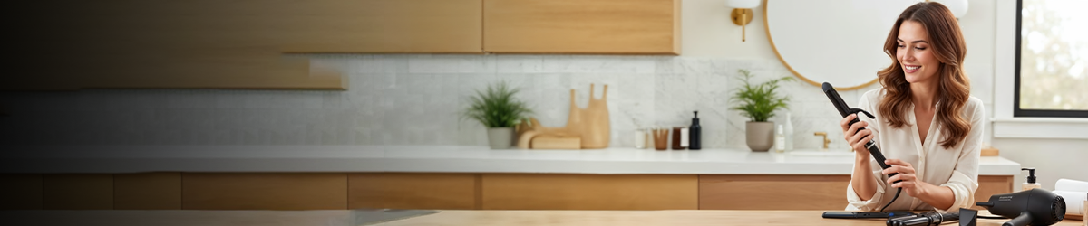 Smiling woman sits at a bathroom counter holding a curling iron, with text advertising curling irons and wands at TED’s Electronics, and a stylish, modern bathroom in the background.